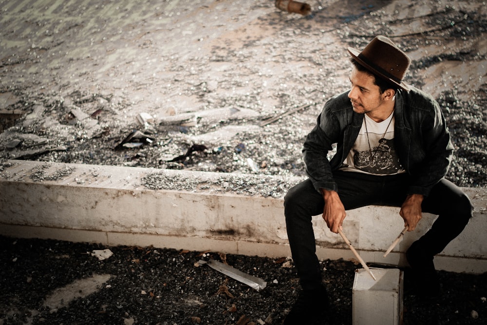 man in black coat sitting on concrete fence holding drumstick