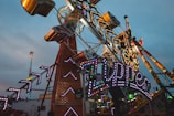 A bustling amusement park scene features a large, colorful ride with illuminated signs and lights. The ride structure is intricate, with angular designs and flashing lights. In the background, a Ferris wheel and other rides add to the vibrant, fun atmosphere.