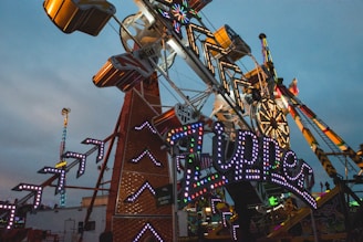 A bustling amusement park scene features a large, colorful ride with illuminated signs and lights. The ride structure is intricate, with angular designs and flashing lights. In the background, a Ferris wheel and other rides add to the vibrant, fun atmosphere.