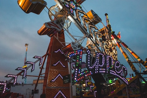 A bustling amusement park scene features a large, colorful ride with illuminated signs and lights. The ride structure is intricate, with angular designs and flashing lights. In the background, a Ferris wheel and other rides add to the vibrant, fun atmosphere.
