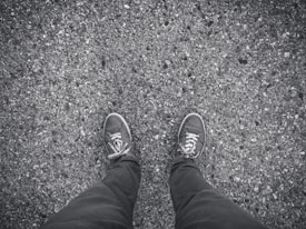 The image depicts a pair of feet wearing casual sneakers on a rough, textured asphalt surface. The ground is speckled with small stones, adding a gritty appearance.