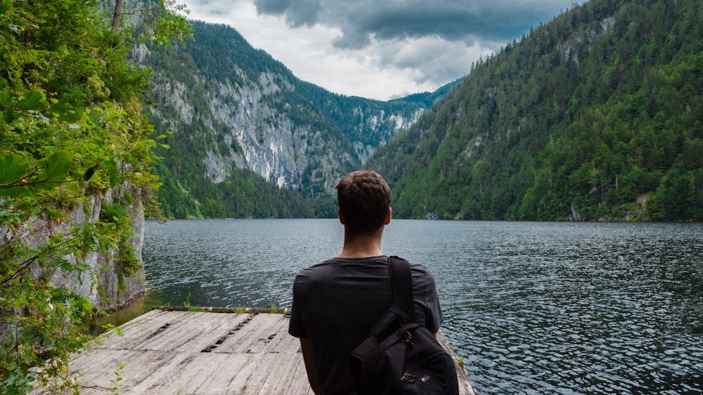 Homme debout sur une planche de bois brun près d’un plan d’eau entouré de montagnes