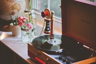 A cozy retro scene with a record player on a windowsill, surrounded by potted plants and a vintage globular lamp. Pink flowers in a glass jar add a touch of color. A vintage-style telephone with a red, white, and blue design is on the side.