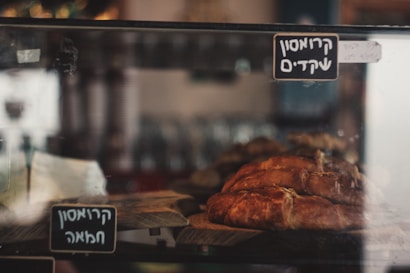 A display case containing freshly baked bread, with golden brown crusts, is shown in a bakery setting. The breads are placed on wooden boards, and there are labels with text written in a non-Latin script attached to the glass display.