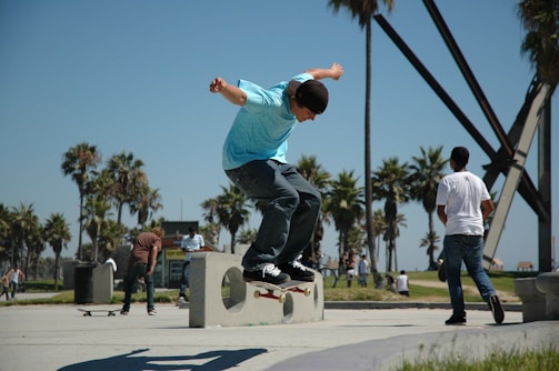 A person performs a skateboard trick on a concrete structure in a sunny outdoor park setting. The area is surrounded by palm trees, with other people walking and skating in the background. The sky is clear and the surroundings suggest a vibrant, energetic atmosphere.