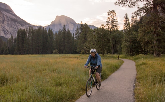 A person is cycling along a paved path through a lush green meadow with towering pine trees in the background. The scene is set against a mountainous landscape with a prominent granite peak visible in the distance.