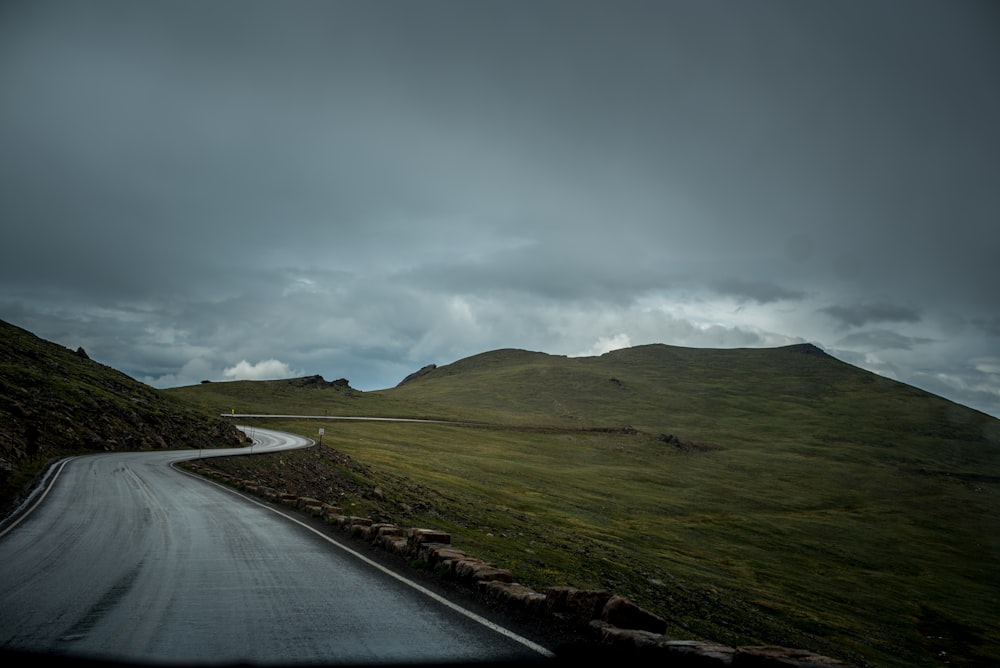 photo of gray concrete road between green mountain and green grass field under cloudy sky