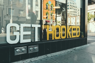 A storefront window displays large white and yellow letters spelling 'GET HOOKED'. Reflections of city buildings and pedestrians can be seen on the glass, along with pineapples placed at each end of the display. The street scene suggests a vibrant urban environment.