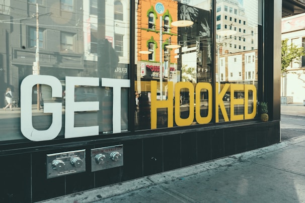 A storefront window displays large white and yellow letters spelling 'GET HOOKED'. Reflections of city buildings and pedestrians can be seen on the glass, along with pineapples placed at each end of the display. The street scene suggests a vibrant urban environment.