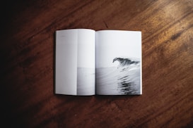 An open book lies on a wooden surface with a black-and-white image of a large ocean wave on one of its pages.