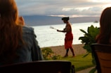A person wearing a traditional garment and a headpiece is performing a dance on a lawn near the beach. There is a scenic ocean view in the background with mountains and a cloudy sky.