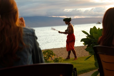 A person wearing a traditional garment and a headpiece is performing a dance on a lawn near the beach. There is a scenic ocean view in the background with mountains and a cloudy sky.