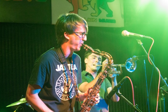 A musician plays the saxophone on stage, wearing glasses and a dark t-shirt with a graphic design. Another musician in the background plays the trumpet. Microphones and a drum kit are also visible under colorful stage lighting.