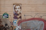 A wall with urban graffiti and street art. There is a detailed portrait of a man painted on a rectangular section of the wall. To the left, a street sign reads 'Strada Arthur Verona'. Surrounding the portrait are various graffiti tags and colorful patterns. The overall aesthetic is gritty and artistic.