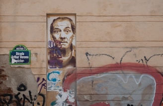 A wall with urban graffiti and street art. There is a detailed portrait of a man painted on a rectangular section of the wall. To the left, a street sign reads 'Strada Arthur Verona'. Surrounding the portrait are various graffiti tags and colorful patterns. The overall aesthetic is gritty and artistic.