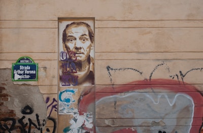 A wall with urban graffiti and street art. There is a detailed portrait of a man painted on a rectangular section of the wall. To the left, a street sign reads 'Strada Arthur Verona'. Surrounding the portrait are various graffiti tags and colorful patterns. The overall aesthetic is gritty and artistic.