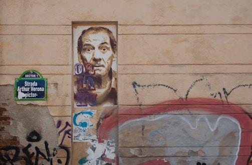 A wall with urban graffiti and street art. There is a detailed portrait of a man painted on a rectangular section of the wall. To the left, a street sign reads 'Strada Arthur Verona'. Surrounding the portrait are various graffiti tags and colorful patterns. The overall aesthetic is gritty and artistic.