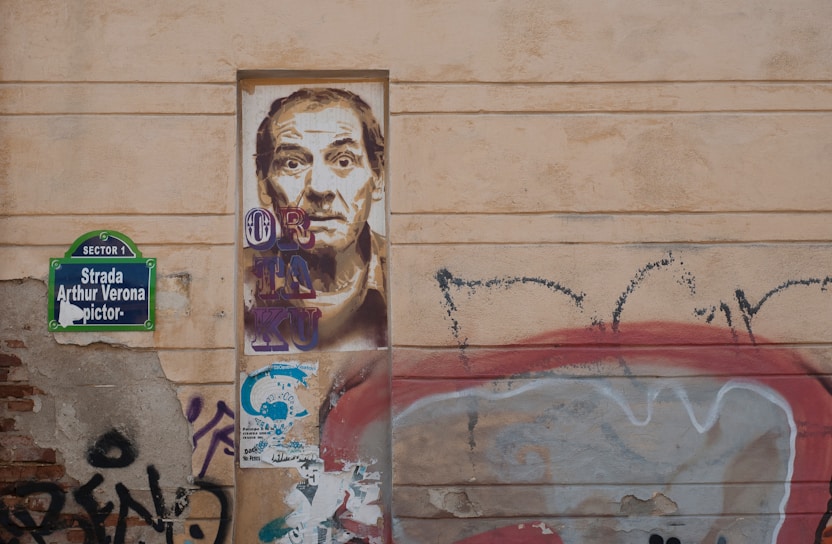 A wall with urban graffiti and street art. There is a detailed portrait of a man painted on a rectangular section of the wall. To the left, a street sign reads 'Strada Arthur Verona'. Surrounding the portrait are various graffiti tags and colorful patterns. The overall aesthetic is gritty and artistic.