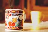 A close-up of a beautifully decorated ceramic mug with intricate floral and bird designs in vibrant colors, placed on a tabletop with another blurred mug in the background. The setting is bathed in warm, natural light creating a cozy atmosphere.