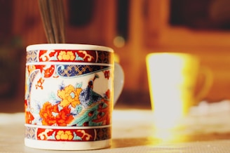 A close-up of a beautifully decorated ceramic mug with intricate floral and bird designs in vibrant colors, placed on a tabletop with another blurred mug in the background. The setting is bathed in warm, natural light creating a cozy atmosphere.
