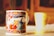A close-up of a beautifully decorated ceramic mug with intricate floral and bird designs in vibrant colors, placed on a tabletop with another blurred mug in the background. The setting is bathed in warm, natural light creating a cozy atmosphere.