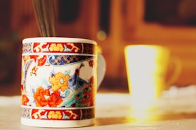 A close-up of a beautifully decorated ceramic mug with intricate floral and bird designs in vibrant colors, placed on a tabletop with another blurred mug in the background. The setting is bathed in warm, natural light creating a cozy atmosphere.