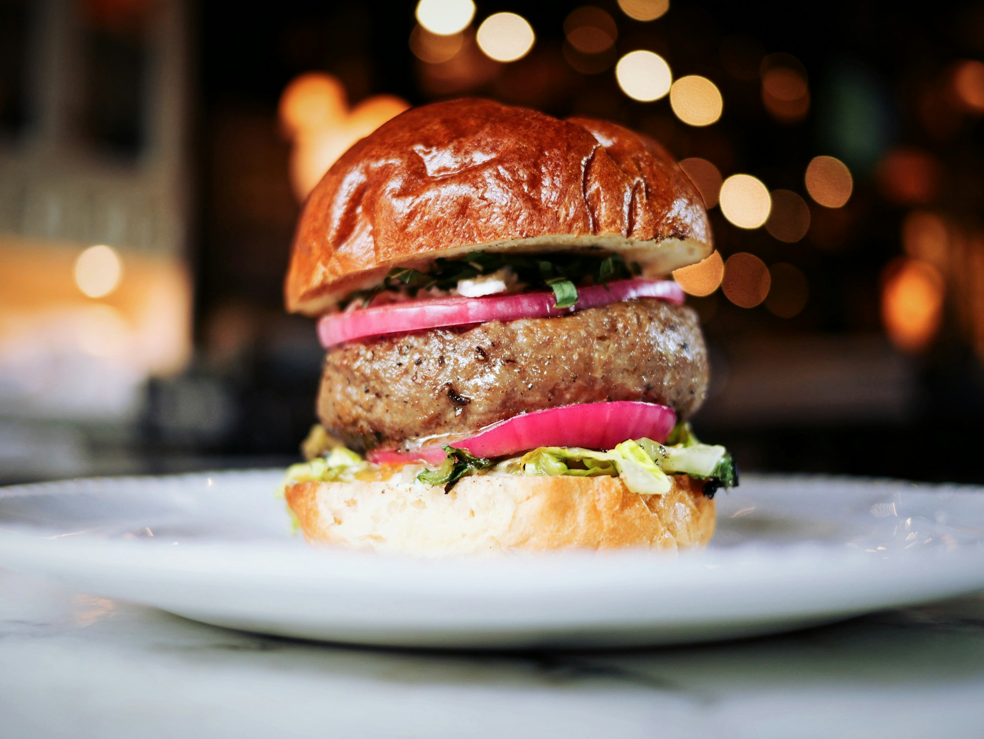 A close-up of a gourmet burger with melted cheese and fresh lettuce, served on a rustic wooden table with soft pastel tones in the background.