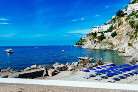 A stunning coast with clear blue water, rocky cliffs adorned with white houses and lush greenery. Several empty beach chairs with blue umbrellas line the shore. Boats float on the calm sea, with sunny and clear skies above.