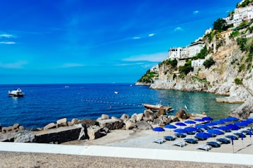A stunning coast with clear blue water, rocky cliffs adorned with white houses and lush greenery. Several empty beach chairs with blue umbrellas line the shore. Boats float on the calm sea, with sunny and clear skies above.
