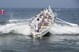 A team of rowers is actively engaged in a rowing competition on the ocean, navigating through waves with their boat's oars extended outwards. The athletes are wearing matching attire as they work in unison against the backdrop of a clear blue sky and ocean. A bright buoy is visible in the water nearby.
