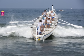 A team of rowers is actively engaged in a rowing competition on the ocean, navigating through waves with their boat's oars extended outwards. The athletes are wearing matching attire as they work in unison against the backdrop of a clear blue sky and ocean. A bright buoy is visible in the water nearby.