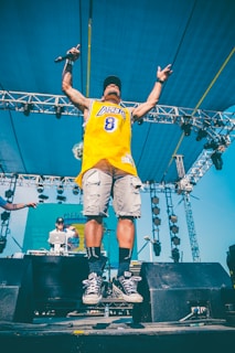 A person wearing a yellow sports jersey and denim shorts is performing on stage, holding a microphone and captured mid-air as they jump. Behind them, a DJ stands at a console with equipment set up on stage. The background features a blue sky and a metal framework with lighting fixtures.