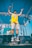 A person wearing a yellow sports jersey and denim shorts is performing on stage, holding a microphone and captured mid-air as they jump. Behind them, a DJ stands at a console with equipment set up on stage. The background features a blue sky and a metal framework with lighting fixtures.