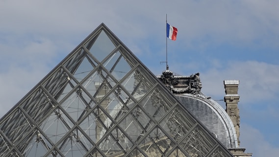 A modern glass pyramid structure juxtaposed with a classical stone building. A French flag is prominently displayed, fluttering above an ornate architectural element.
