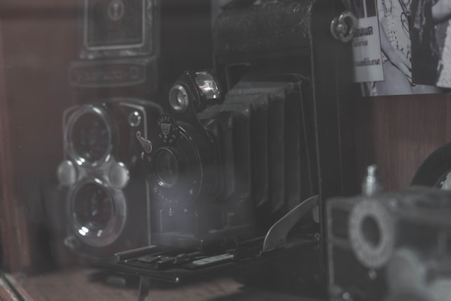 A collection of vintage cameras, mostly black with metal accents and aged textures, is displayed alongside faded black-and-white photographs. The cameras have intricate designs, with various knobs and lenses, evoking a nostalgic feel.
