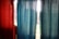 Two colored curtains, one in a textured red material and the other in a smooth blue fabric, hang side by side. Sunlight streams through the window behind them, creating a glow around the curtains and highlighting the textures.
