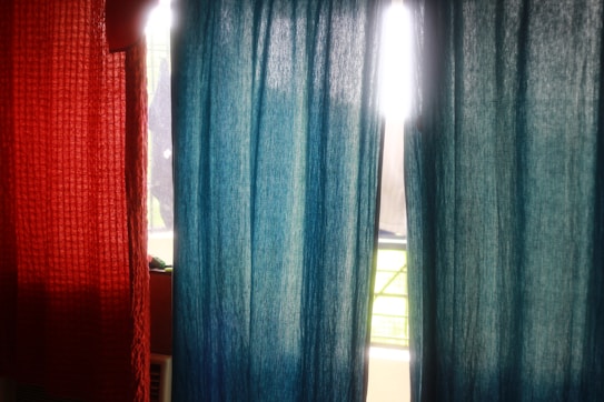Two colored curtains, one in a textured red material and the other in a smooth blue fabric, hang side by side. Sunlight streams through the window behind them, creating a glow around the curtains and highlighting the textures.