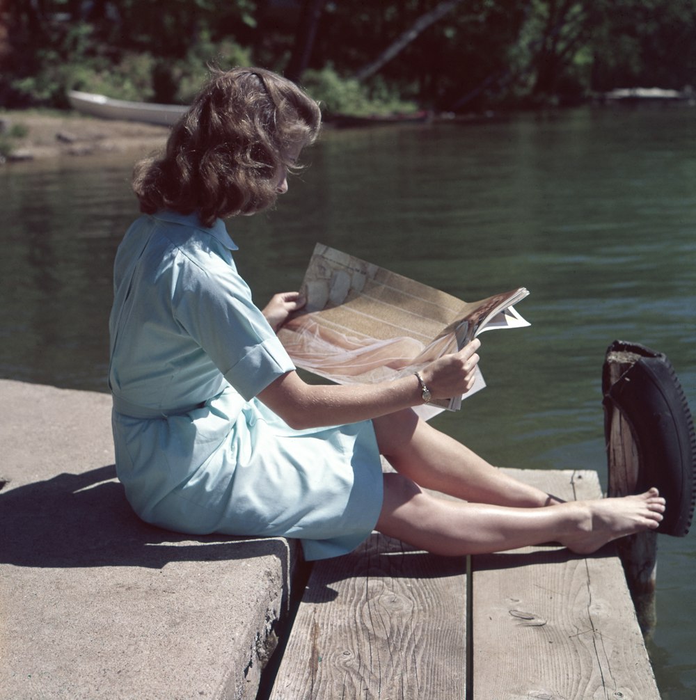 femme portant une robe bleue lisant un magazine près d’un plan d’eau pendant la journée
