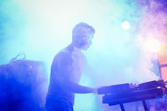 A person is standing at a keyboard or synthesizer with colorful, thick stage smoke swirling around. The scene is bathed in vibrant blue, purple, and pink lighting, creating a dynamic and atmospheric environment.