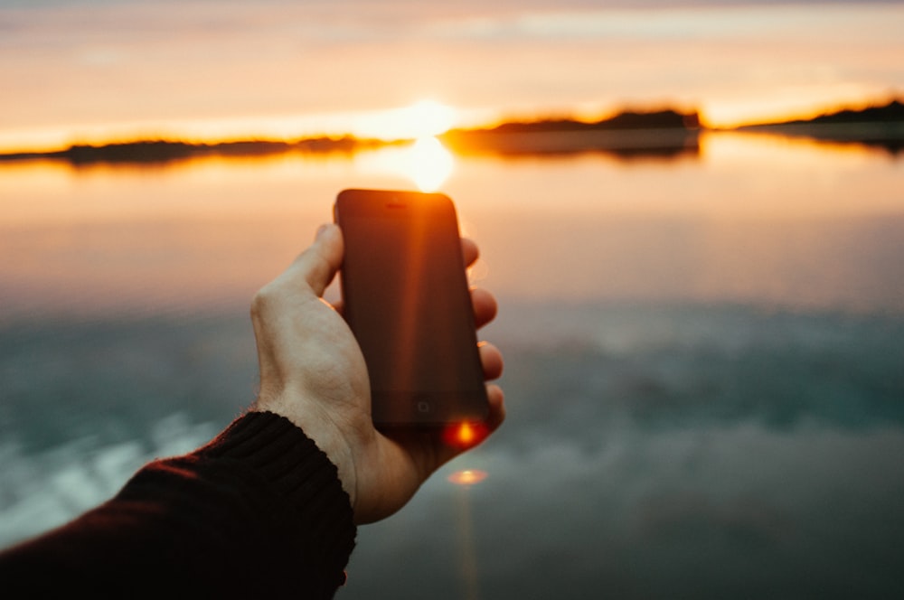 Person mit schwarzem iPhone in der Nähe des Sees während des Sonnenuntergangs