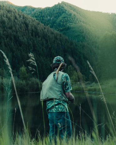 A close-up of a rugged hunter wearing a camo hat, standing by a misty pond at dawn.