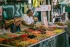 A bustling outdoor market stall with a variety of colorful candies neatly arranged in bins. A man in a white shirt stands behind the counter engaged in conversation with a customer. The background features vivid graffiti art on the wall, adding an urban and vibrant atmosphere to the scene. Numbers indicating prices are prominently displayed on signs.