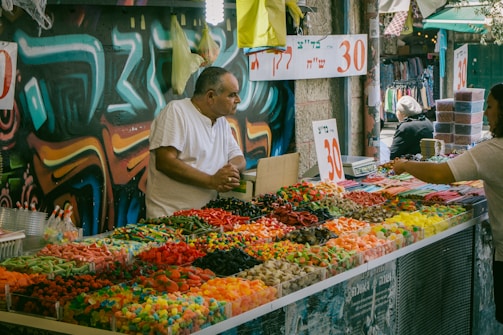 A bustling outdoor market stall with a variety of colorful candies neatly arranged in bins. A man in a white shirt stands behind the counter engaged in conversation with a customer. The background features vivid graffiti art on the wall, adding an urban and vibrant atmosphere to the scene. Numbers indicating prices are prominently displayed on signs.