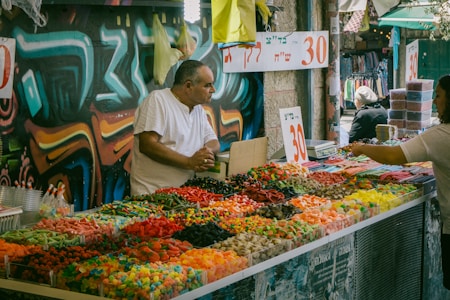 A bustling outdoor market stall with a variety of colorful candies neatly arranged in bins. A man in a white shirt stands behind the counter engaged in conversation with a customer. The background features vivid graffiti art on the wall, adding an urban and vibrant atmosphere to the scene. Numbers indicating prices are prominently displayed on signs.