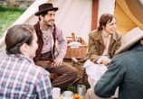 A group of people in vintage clothing sit together outside a canvas tent. The setting has a rustic and historical feel, with a man wearing a brown hat and vest and a woman beside him in a coat. They are engaging in conversation around a picnic table, with a wicker basket filled with dishes and utensils.