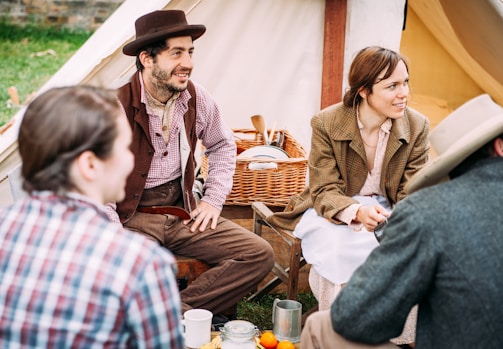 A group of people in vintage clothing sit together outside a canvas tent. The setting has a rustic and historical feel, with a man wearing a brown hat and vest and a woman beside him in a coat. They are engaging in conversation around a picnic table, with a wicker basket filled with dishes and utensils.