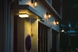 An exterior wall of a building features several lit wall-mounted lantern-style lights. There is a rectangular fluorescent lamp at the bottom left corner above a dark decorative wall piece. The scene is set during the evening or nighttime, with shadows and a tree canopy visible in the background.