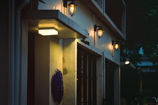 An exterior wall of a building features several lit wall-mounted lantern-style lights. There is a rectangular fluorescent lamp at the bottom left corner above a dark decorative wall piece. The scene is set during the evening or nighttime, with shadows and a tree canopy visible in the background.