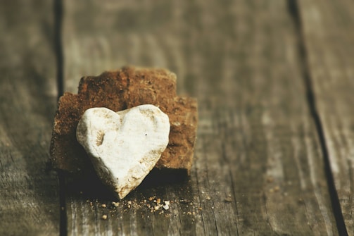 A heart-shaped white stone rests against a rough brown rock on a weathered wooden surface, creating a rustic and natural appearance.
