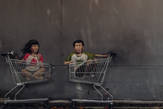 A mural painting depicts two children sitting in shopping carts, one girl and one boy, with joyful expressions. The artwork is on a concrete wall and includes vibrant colors on their clothing, enhancing the playful scene.